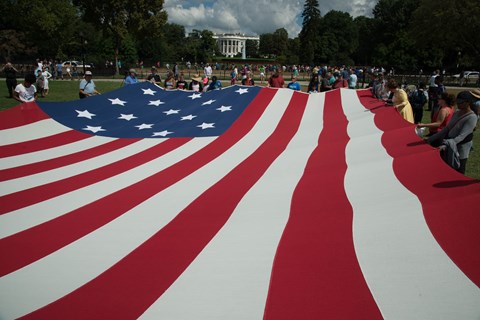 A crowd of people holding a large Star-Spangled Banner replica flag in front of the White House