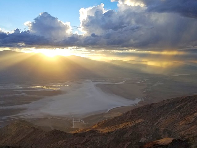 Shafts of light stream through clouds over a dark mountain range and swirling white salt flats.