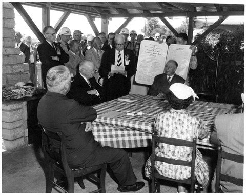 A 1950s photo depicts dignitaries honoring a distinguished old man at a picnic shelter.