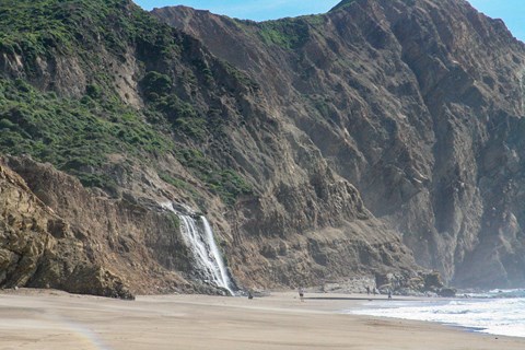 White water cascading over grass-topped coastal bluffs to a sandy ocean beach with four people below