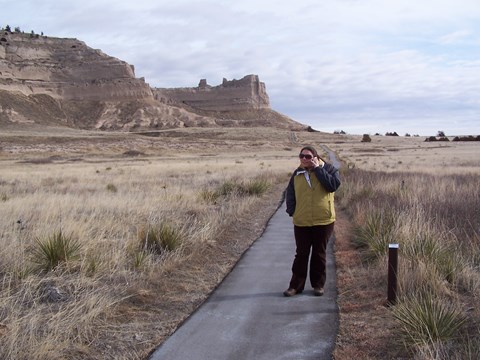 A woman stands on a paved trail with a cellular phone to her ear and a view of sandstone bluffs.