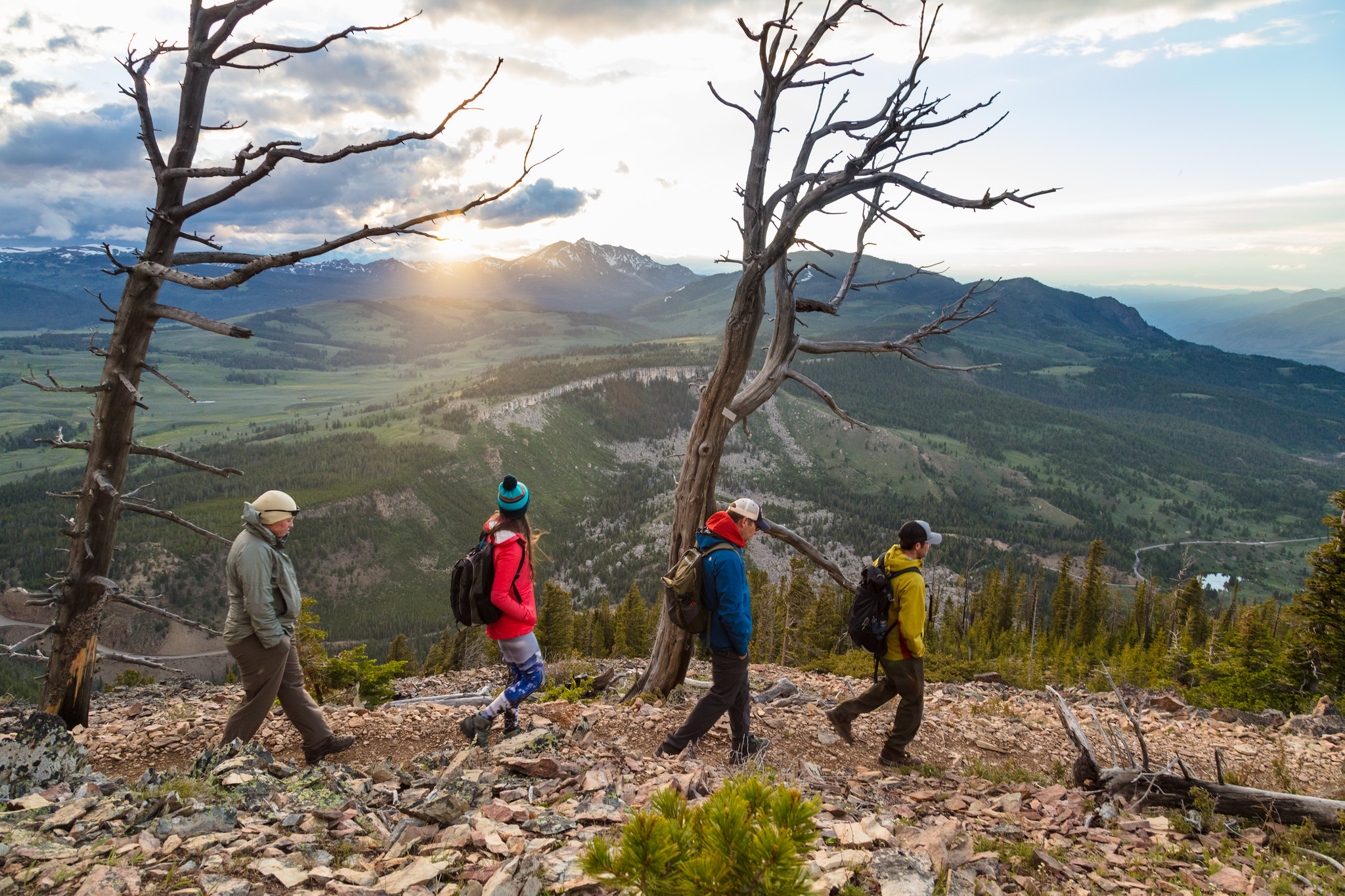 Four people hike across a talus slope with the sun setting behind a mountain in the distance.