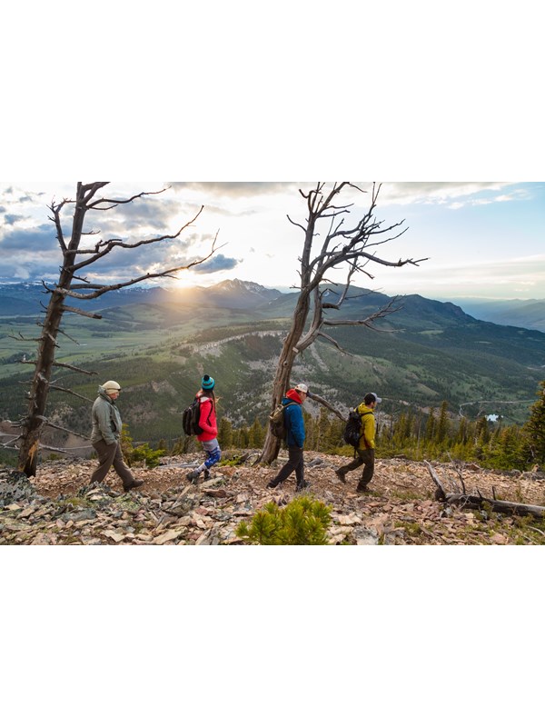 Four people hike across a talus slope with the sun setting behind a mountain in the distance.