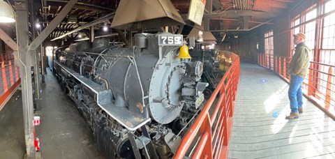 Visitor wearing mask while standing on elevated platform in roundhouse, looking at a train