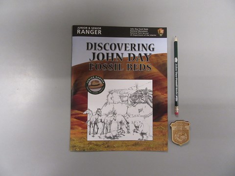 Book with text "Discovering John Day Fossil Beds" next to a wooden Junior Ranger badge and pencil