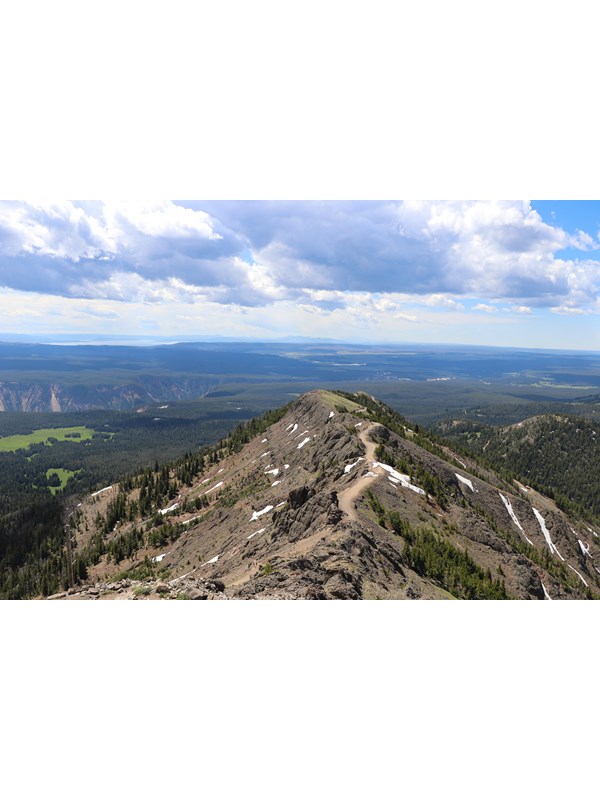 A trail crosses a ridgeline above forests and meadows.