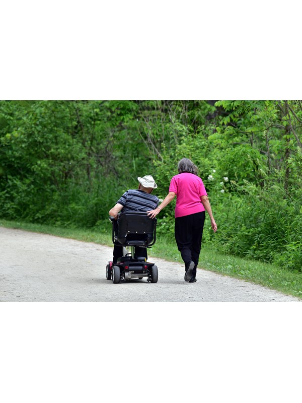 A woman walks beside a man on a flat trail, resting a hand on the back of his motorized wheelchair.