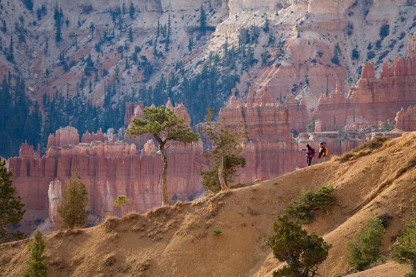 Two hikers descend down a trail from right to left with red rock formations in the background