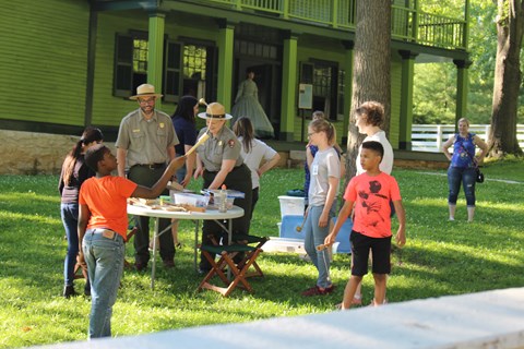 Two park rangers at a table standing alongside five students with a green house in the background.