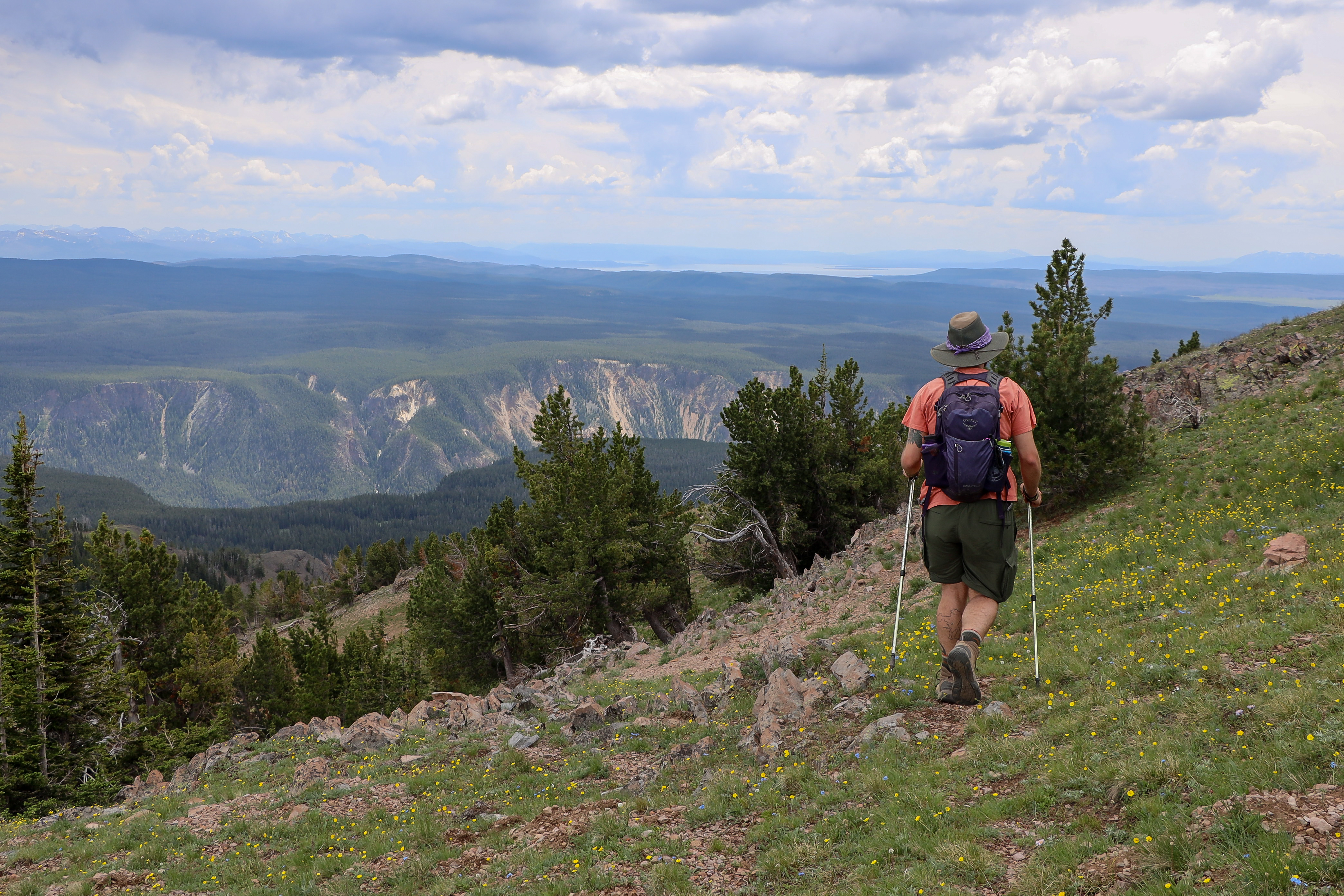A hiker walks on a mountain slope with a canyon and lake seen in the distance.