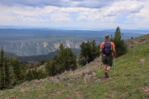 A hiker walks on a mountain slope with a canyon and lake seen in the distance.