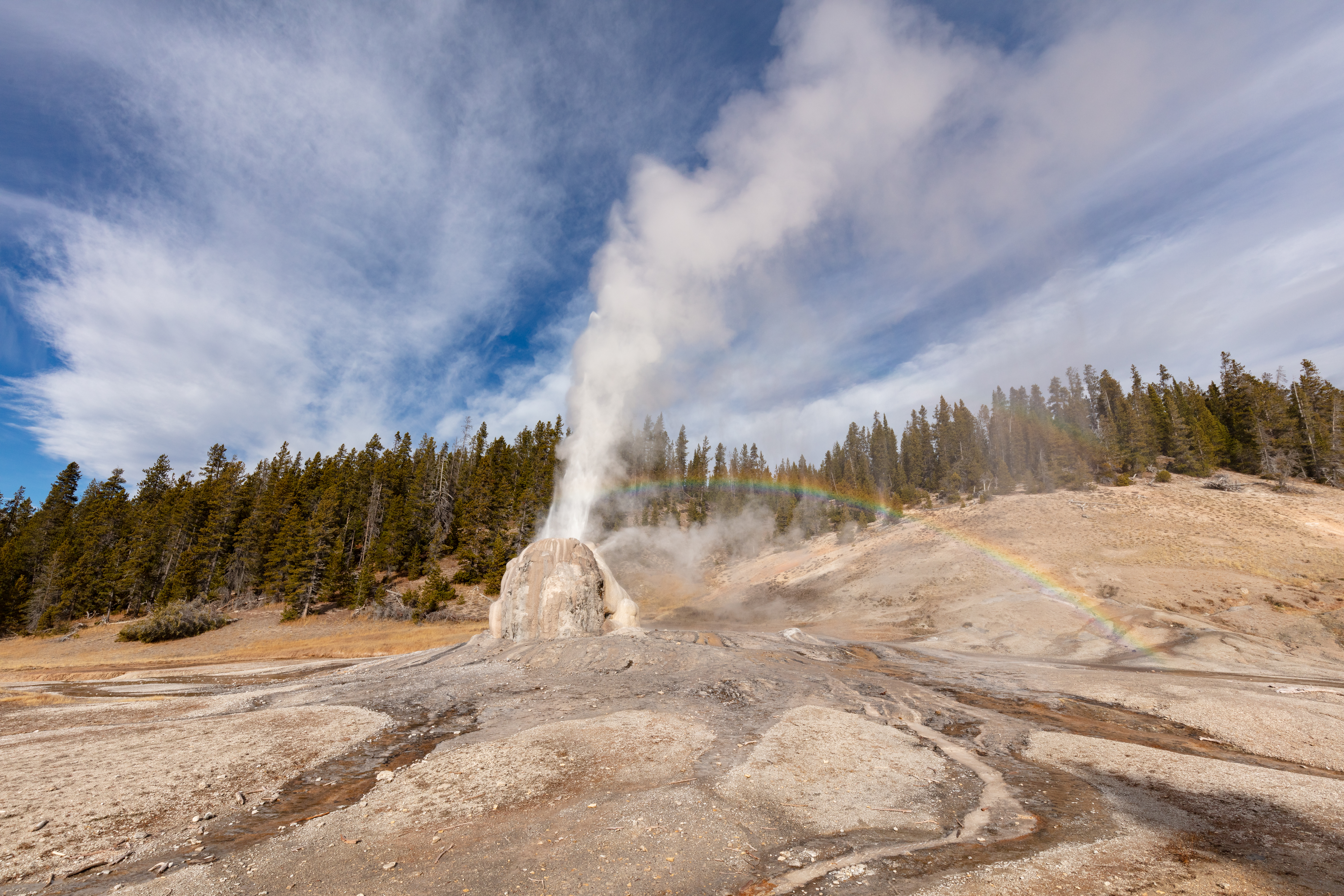 Steam and water erupt from the tan cone of Lone Star Geyser. A rainbow appears in the mist.