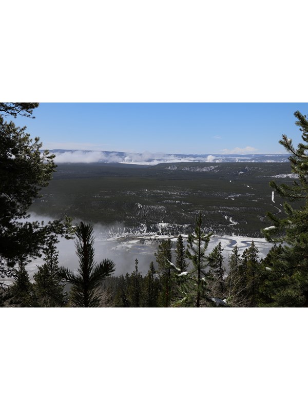 Steam from geyser basins dot a forested landscape as viewed from a mountain summit.