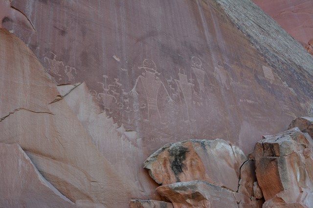 Rock markings carved into flat red rock showing human-like figures with trapezoidal bodies