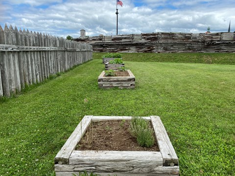 3 raised garden beds in a grassy area in front of the fort wall and inside the picket fence.