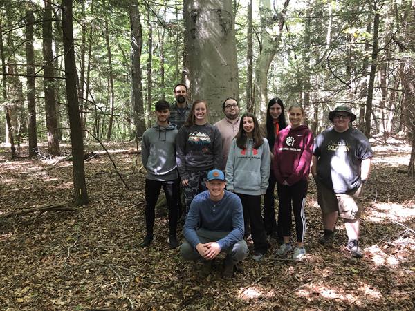 4 young women, 4 young men, and an older man posing and smiling in front of a large tree in a forest