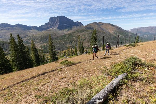 Two backpackers walk along a trail with a mountain peak in the background.