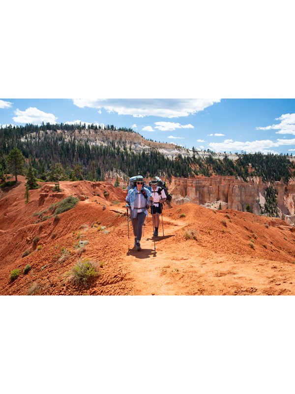 Two people walk a trail of red earth wearing gear on their backs.