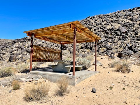 A shaded picnic pavilion with table with a lava rock escarpment in the background.
