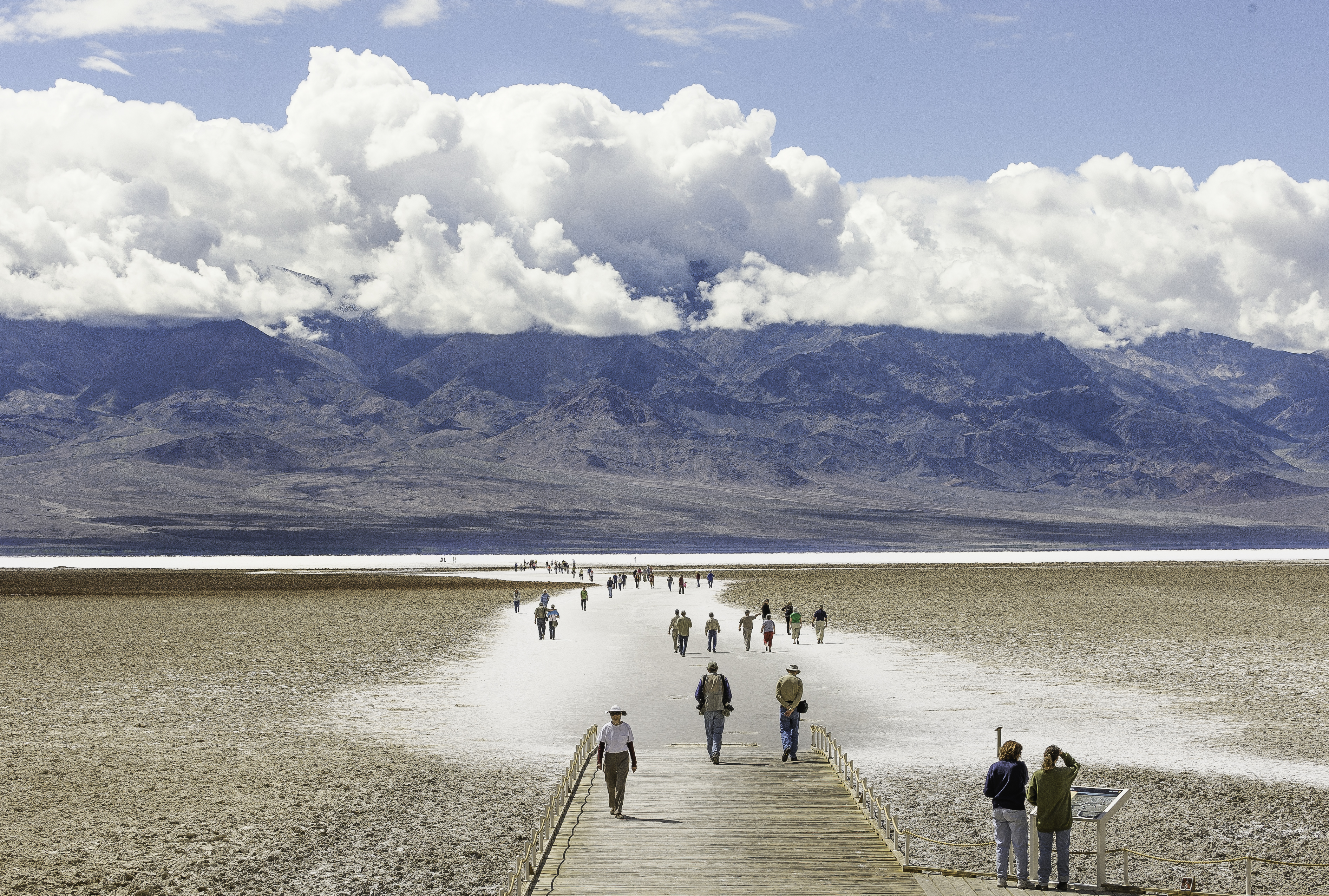 The image shows a broad, arid landscape with a wooden path leading deep into a salt flat.