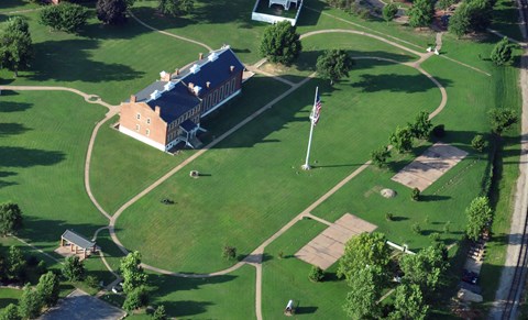 A grassy field with pathway that loops around a flagpole with radiating paths and brick buildings.