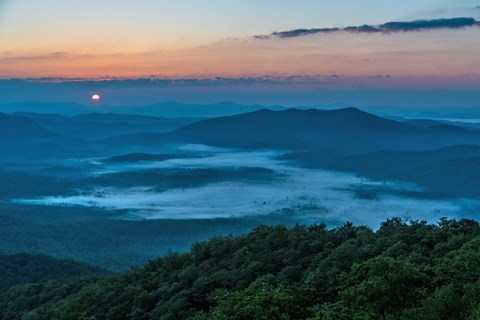 The sun rises above mist filled valleys surrounded by mountains.