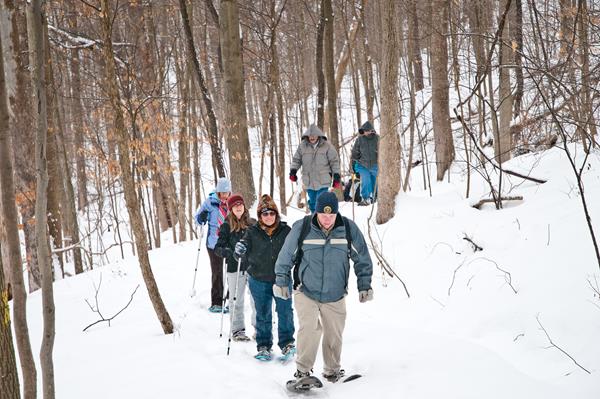A group of people in winter hats snowshoes along a snow-covered trail in the forest.