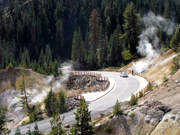 A road lined by wooden guardrails curves through a hydrothermal area of barren, steaming ground.
