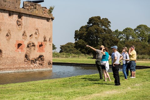 A park ranger pointing at a battle-damaged brick wall with a crowd behind.