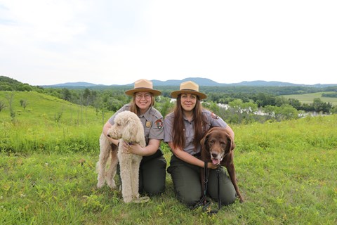 Two women in park ranger uniforms kneel next to two dogs on leash.