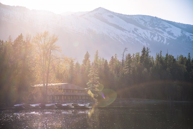Sunlit Apgar Village lodge building above a row of small boats docked on Lake McDonald, with a snowy mountain behind