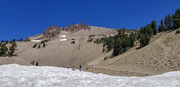 People play on a field of snow below a hiking trail at the base of large volcanic dome.