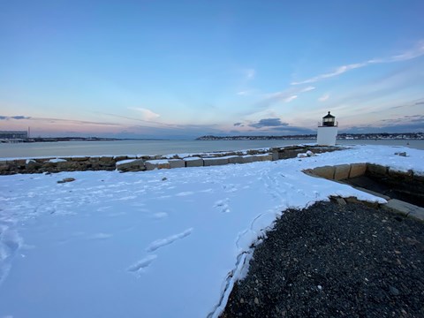 Light blue morning light over snowy wharf pathway with white lighthouse in distance.