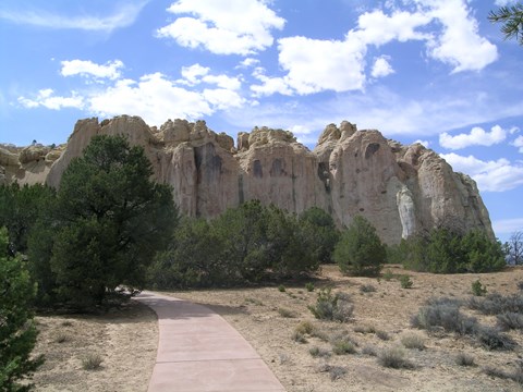 A trail winds through juniper trees towards the base of a large cliff.