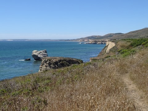 Beige-colored coastal bluffs on the right line a bay with a guano-covered seastack on the left.