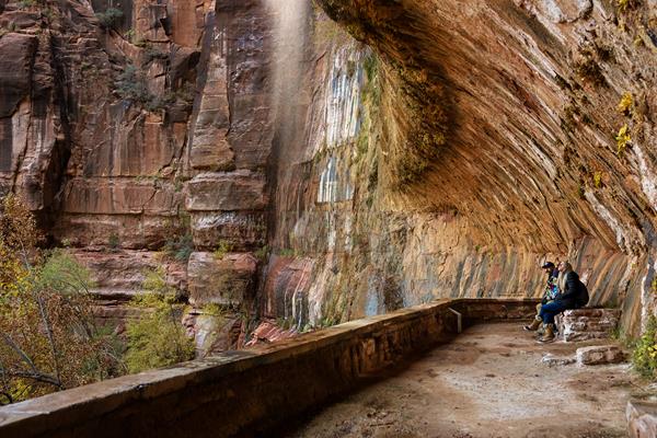 Two people sit in a rock alcove, with water dripping off the overhang in front of them.