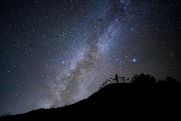 A silhouetted figure stands at a fenced overlook with the milky way arching above