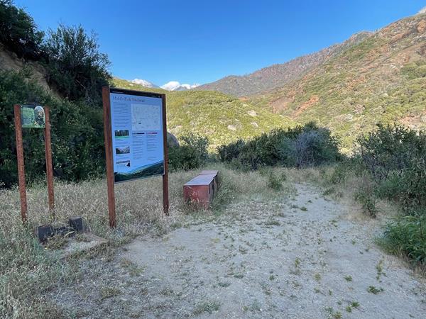 A trailhead sign and bench are located at the beginning of a dirt trail with a hill in background.