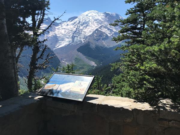 A wayside panel attached to a low rock wall overlooks a deep glacier valley and glaciated mountain.