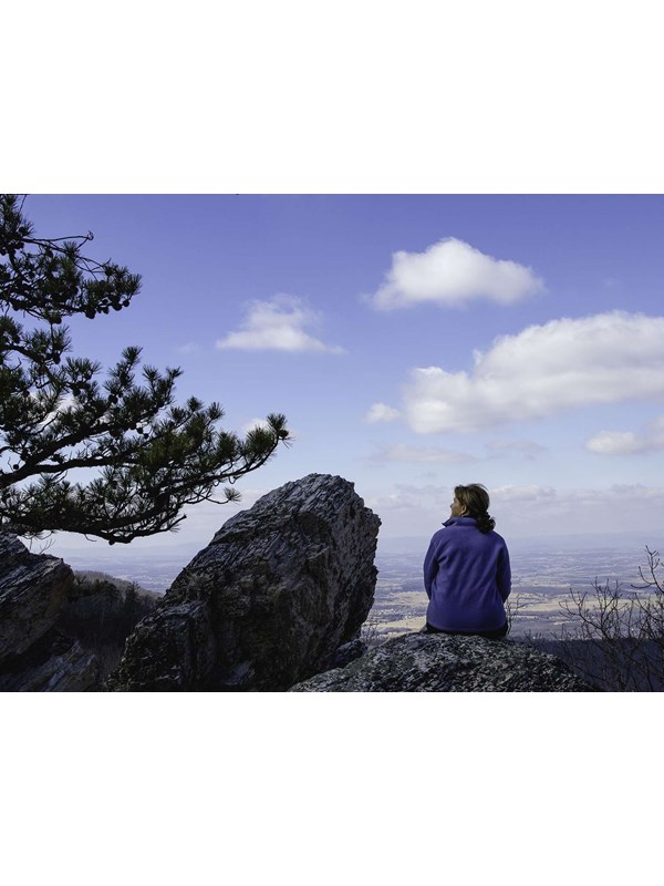 A color photograph of a woman sitting on a rock atop a mountain looking to the side.