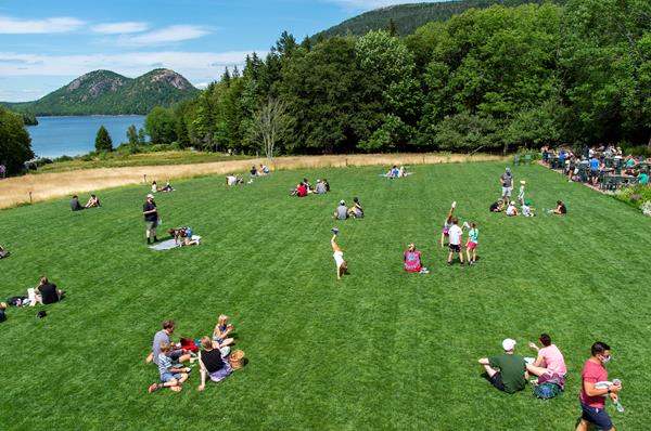 Groups of people sit on a mowed lawn in front of a lake and distant mountains