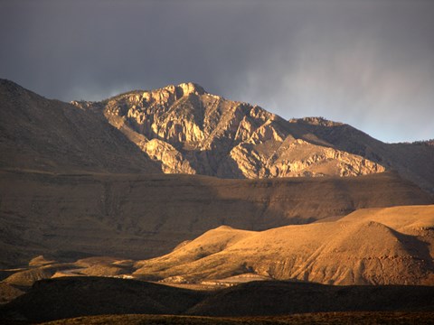 A desert mountain peak is lit by the setting sun as it rises above the landscape around it.