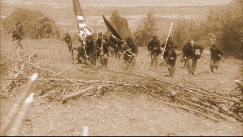 A sepia toned still image of reenactors running up hill with flags and guns.