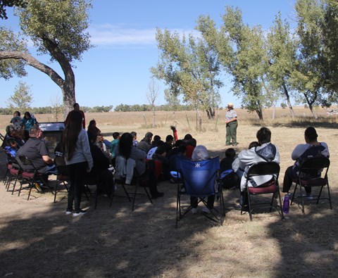 A ranger stands in an open area speaking to a group of about 20 seated people.