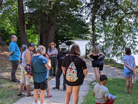 Park ranger standing in front of a group of visitors in the shade with a pond behind them.n