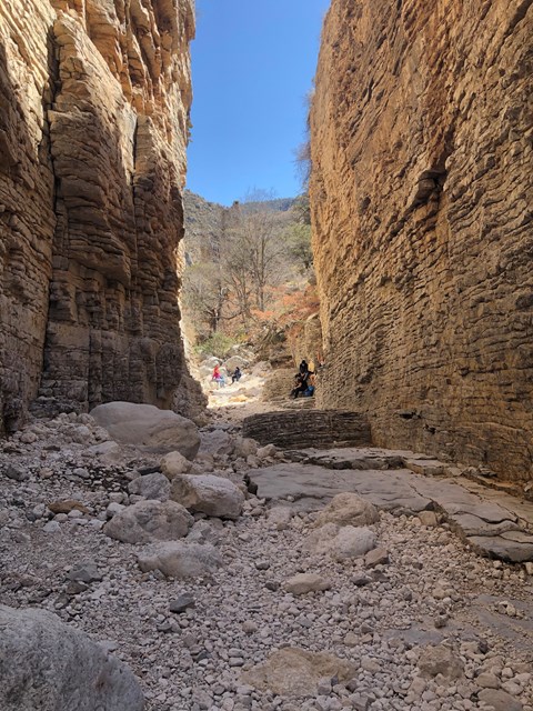 Groups of people in the sunlight at the end of a narrow tall canyon.