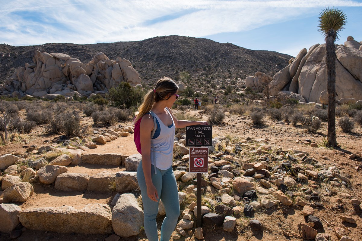 A hiker next to a sign for Ryan Mountain by a dirt and rock trail heading towards a mountain.