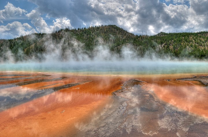 Steam rises off of a large, blue hot spring surrounded by yellow, orange, and brown microbial mats.