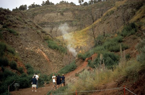 A small group of people stand at the base of a valley, watching smoke rise from the ground.
