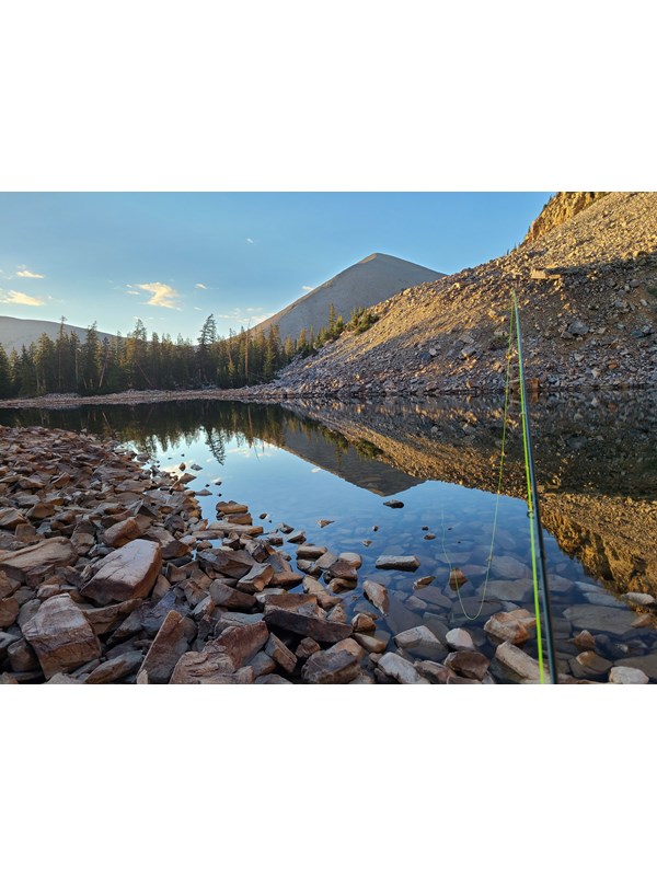 A reflective lake surrounded by rocks, with a green fishing rod emerging from the viewer.
